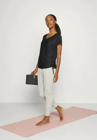 Crna kratka majica s V-izrezom i bočnim vezanjem, u kombinaciji sa svijetlosivim uskim hlačama. Model hoda bos na ružičastom jogamat.