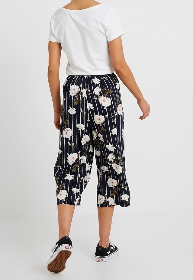 Marina beskurna byxor med vitt blommönster och vertikala ränder, med elastisk midja och avslappnad passform, tillsammans med svarta sneakers.