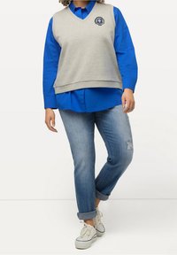 Ljust grå ärmlös tröja med v-ringning och logotyp, lager på lager över en blå långärmad skjorta, kombinerad med slitna blå jeans och vita sneakers.