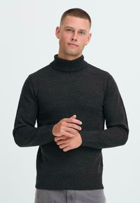 Jonge man met kort haar, gekleed in een zwart coltrik, en grijze broek, staande met zijn handen gevouwd tegen een witte achtergrond.