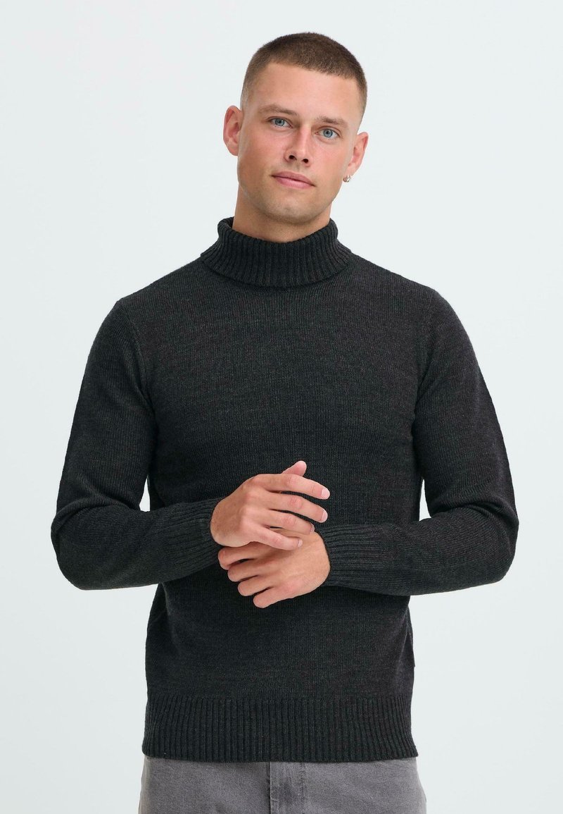 Jonge man met kort haar, gekleed in een zwart coltrik, en grijze broek, staande met zijn handen gevouwd tegen een witte achtergrond.