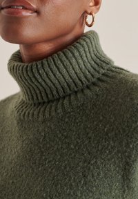 Olivgrön polotröja i texturerad stickad väv, med en ribbad krage. Närbild visar mjukhet och detaljerad sömnad.