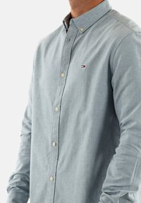 Camisa abotonada de tela azul clara, con cuello con solapa, botones dorados y un pequeño logo en el pecho. Textura suave.