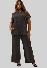 Vrouw in een bijpassende donkere textuur met korte mouwen en wijde broek, staand met een hand in haar zak en zwarte hakken.