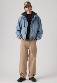 Τζιν μπουφάν με κουκούλα, ανοιχτό μπλε χρώμα, και ελαστικά μανίκια; μαύρο μπλουζάκι από κάτω, καφέ παντελόνι, και μαύρα αθλητικά παπούτσια; καπέλο παραλλαγής.