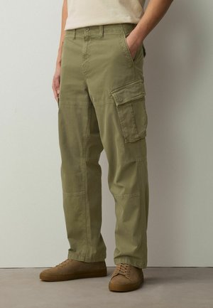 Mann trägt olivgrüne Cargohosen und braune Sneakers, steht mit einer Hand in der Tasche vor einer schlichten Wand.