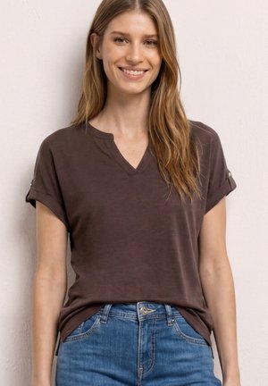 Eine lächelnde Frau trägt ein dunkelbraunes Kurzarmshirt mit V-Ausschnitt, das in blaue Jeans gesteckt ist, und steht vor einer schlichten hellen Wand.
