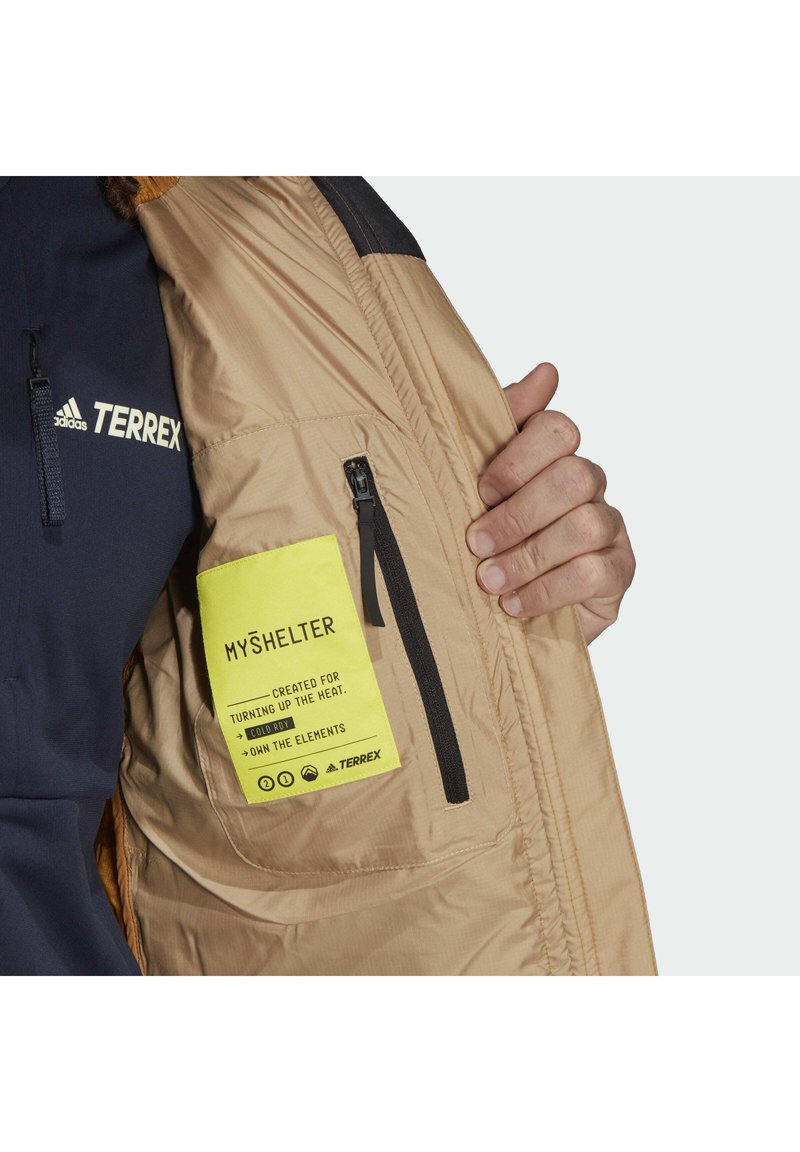 Beige isolierte Jacke mit einem Innenfach und einem Reißverschluss, das ein leuchtend gelbes Etikett enthält. Schwarzer Reißverschluss und teilweise sichtbarer navyfarbener Ärmel.