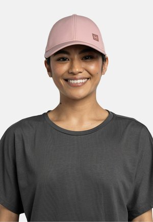 Mujer sonriente con una gorra de béisbol rosa claro y una camiseta gris oscuro holgada sobre un fondo liso y claro.