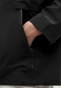Černá lehká bunda s kapsou na zip, elegantní design a hladká textura. Obsahuje jemné logo na rukávu.