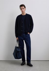 Navy cardigan with button closure and pockets, worn over a dark top. Blue jeans and black shoes complete the outfit. Carrying a dark tote bag.
