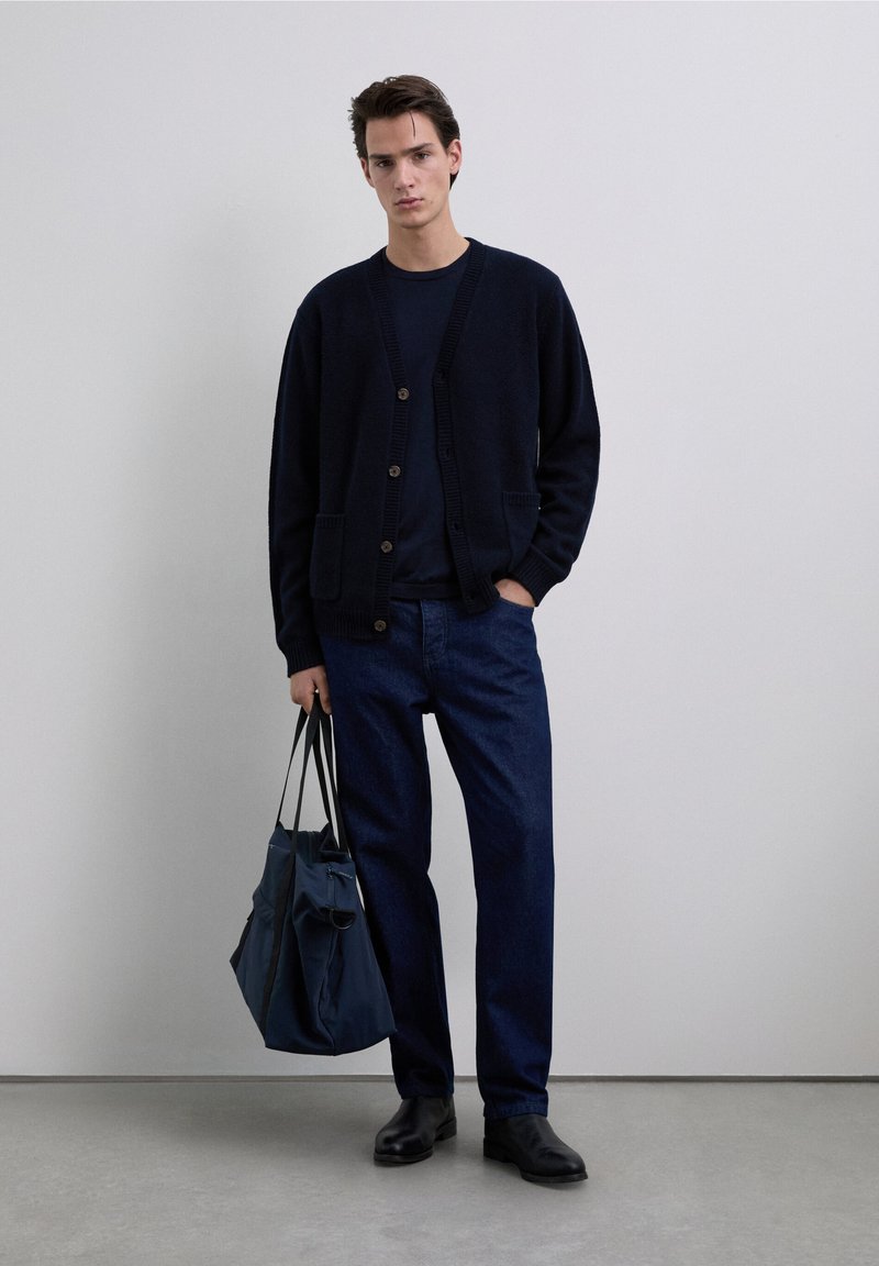 Navy cardigan with button closure and pockets, worn over a dark top. Blue jeans and black shoes complete the outfit. Carrying a dark tote bag.
