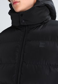 Sort pufferjakke med en høj, polstret krave, der har en central lynlås og trykknapper. Glat, quiltet tekstur med logodetalje.
