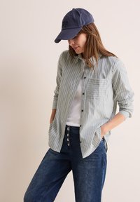 Camicia a righe verde e bianca, con tasca. Abbinata a jeans blu scuro con grandi bottoni. Un cappellino da baseball casual completa il look.