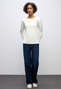 Weißes langärmliges Oberteil mit einem runden Ausschnitt, kombiniert mit dunkelblauen weit geschnittenen Jeans und weißen Sneakers, vor einem neutralen Hintergrund.