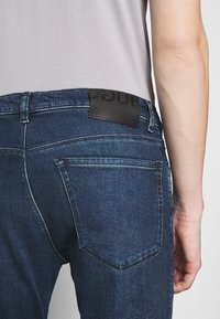 Tmavě modré džíny s přiléhavým střihem, se švy v kontrastní barvě, koženou nášivkou na pase a zadními kapsami s šikmými okraji.