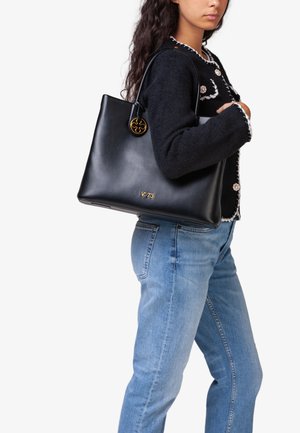 Bolso tote de cuero negro con una textura suave, que presenta un acento circular dorado y un detalle de logo grabado en el lateral.