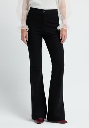 Mujer que lleva pantalones acampanados de talle alto en negro, blusa blanca de manga larga con mangas abullonadas transparentes y zapatos burdeos puntiagudos.