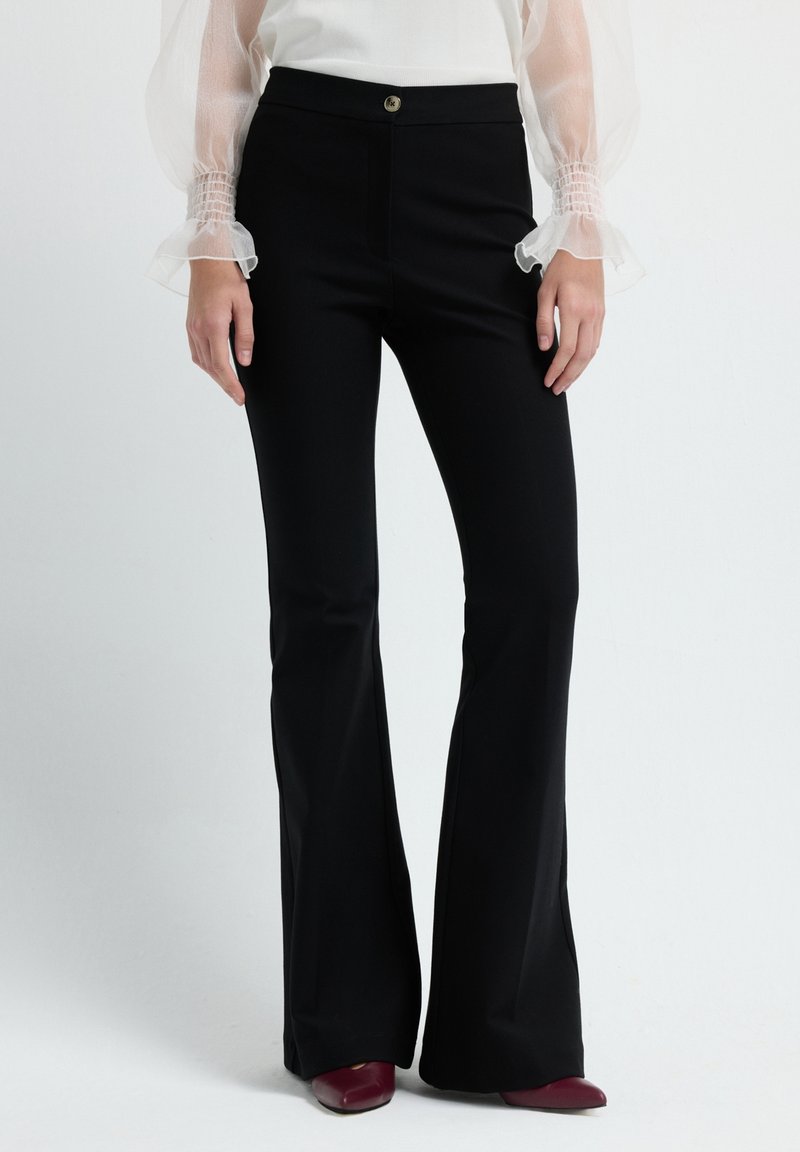 Femme portant un pantalon noir évasé taille haute, un chemisier blanc à manches longues transparentes et bouffantes, et des chaussures bordeaux à bout pointu.