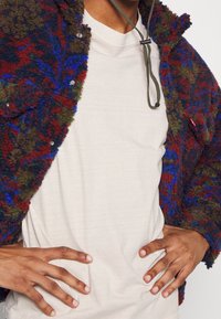 Chlupatý, barevný kabát s vzorem červených, modrých a zelených listů, s patentkami a stahovací šňůrkou kolem krku, přes béžové tričko.