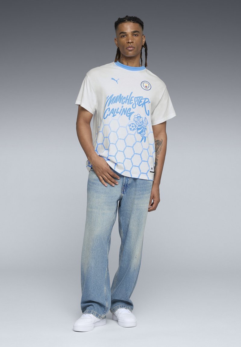 Tricou alb și albastru deschis, cu mânecă scurtă, având un model în plasă de miere, grafică "Manchester Calling" și logo; asortat cu blugi albaștri decolorati.