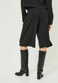 Černé culotte šortky s hladkým povrchem, se bočními rozparky, spárované s vysokými černými koženými botami s akcentem přezky a blokovými podpatky.