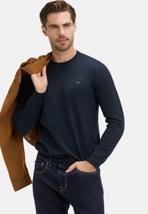 Homme portant un pull marine et un jean, transportant une veste marron sur l'épaule, debout devant un fond blanc.