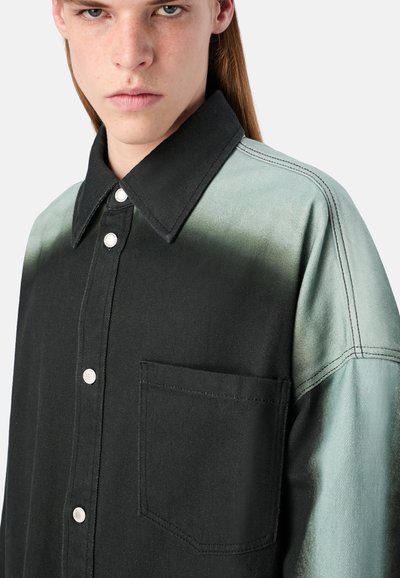 Lähikuva henkilöstä, joka on pukeutunut mustaan ja vaaleanvihreään napitettavaan paitaan, jossa on tasku. Henkilö näyttää vakavalta ja hänellä on pitkät suorat hiukset.