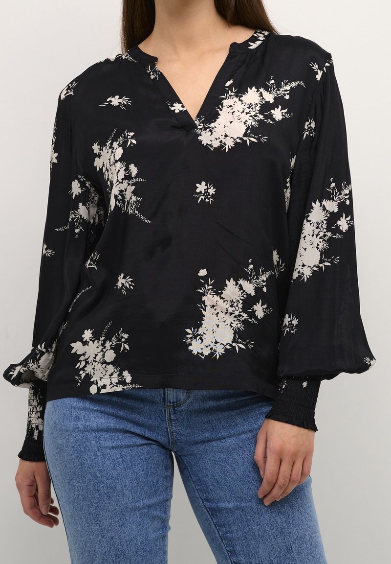 Femme portant un chemisier noir à manches longues avec des imprimés floraux blancs et un jean bleu, debout devant un fond clair uni.