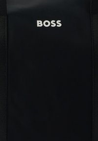 Μαύρο σακίδιο κατασκευασμένο από ανθεκτικό υλικό, με εμφανές λευκό λογότυπο «BOSS» στο μπροστινό μέρος, με υφασματικές λεπτομέρειες και ανθεκτικούς πλευρικούς ιμάντες.
