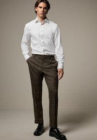 Cămașă albă cu nasturi, pantaloni carouri maro și pantofi negri. Pantalonii au un model discret în carouri. Croială slim fit, cu o textură de țesătură netedă.