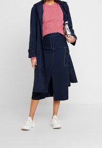 Gonna midi blu navy con spacco sul davanti e tasche, abbinata a un maglione rosa di maglia e un trench blu scuro. Le scarpe da ginnastica bianche completano il look.