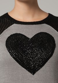 Pull en gris avec des accents en maille noire et un grand design en sequins noirs en forme de cœur au centre. Il possède un col rond et une texture côtelée.