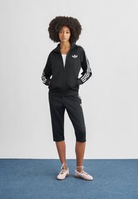 Mustast spordiriietusest komplekt, valgete triipudega, lukuga jakk ja põlveni ulatuvad püksid. Modellil on jalas heledaroosad kingad, millel on ristitriipudega rihmad.