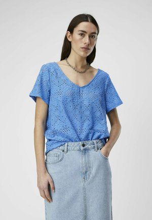 Blå blonde topp med V-hals, korte ermer og blomstermønster, kombinert med et lyst denimskjørt. Modellen har hendene i lommene.