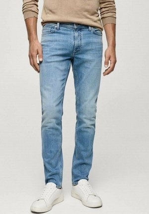 Mann trägt hellblaue Slim-Fit-Jeans und weiße Sneaker, steht vor einem einfarbigen hellen Hintergrund.
