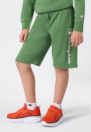 Enfant portant un short et un sweat à capuche Champion verts, avec des chaussettes blanches Champion et des chaussures de sport orange vif, se tenant sur un fond blanc.