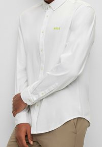 Camicia bianca a maniche lunghe con vestibilità su misura, dotata di logo "BOSS" verde lime, colletto button-down e bottoni verdi chiari. Texture liscia.