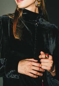 Blusa in velluto nero con collo alto, bottoni arrotondati e maniche a sbuffo. Anello colorato con gioielli e orecchini grandi aggiungono dettagli di stile.