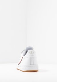 Vit lädersneaker med en strukturerad sula, har ett rött och grönt rand längs sidan och ett präglat logotyp på hälen.