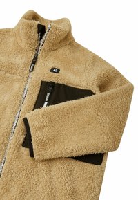 Beige Fleecejacke mit hohem Kragen, Reißverschluss und einer schwarzen Reißverschlusstasche auf der Brust; hat eine weiche, strukturierte Oberfläche.