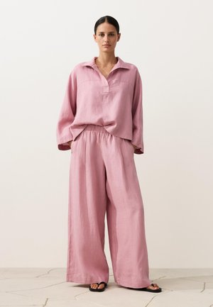 Vrouw draagt een losse, lichtroze linnen top met lange mouwen en bijpassende wijde broek, staand op een stenen vloer tegen een effen achtergrond.