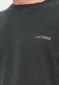 Czarna bawełniana koszulka z okrągłym dekoltem, z białym logo "ARKK" na lewej stronie klatki piersiowej. Miękka tekstura, minimalistyczny design.