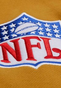Toppa del logo NFL con lettere ricamate rosse su sfondo bianco con contorno blu, su un tessuto giallo senape texturizzato.