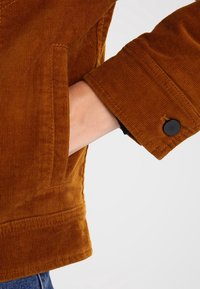 Bruin corduroy jasje met gestructureerde stof, twee voorzakken en een knoop aan de manchet. Zichtbare stiksels en strak ontwerp.