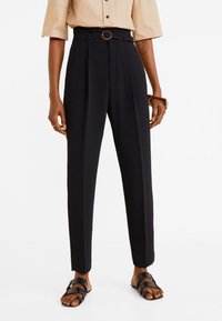 Pantalon noir taille haute avec plis, boucle de ceinture circulaire marron, et coupe fuselée aux jambes, associé à des sandales noires.
