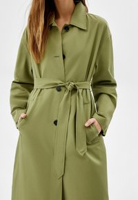 Olivgrön trenchcoat med bälte i midjan, knappstängning och sidofickor. Slät tyg med klassisk krage och långa ärmar.