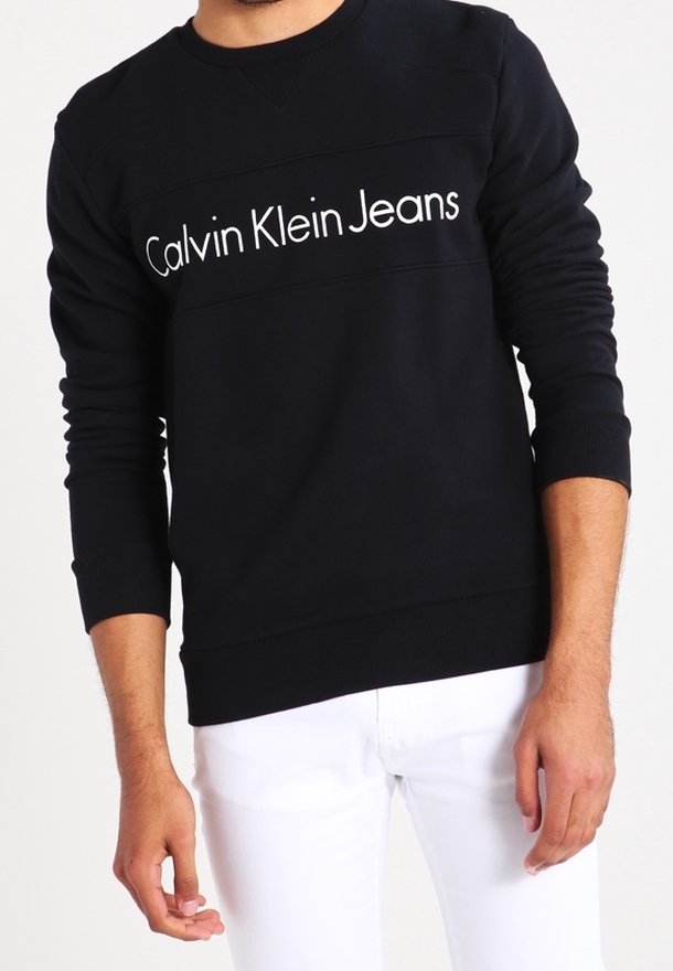 Homme portant un sweat-shirt noir avec le texte « Calvin Klein Jeans » et un pantalon blanc, debout devant un fond clair uni.