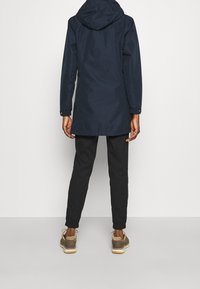 Laivastonsininen vedenpitävä takki, jossa on huppu, pitkät hihat ja säädettävät hihansuut. Yhdistetty mustiin kapeneviin housuihin ja ruskeisiin tennareihin.