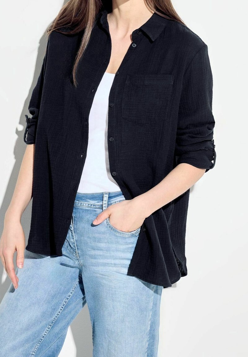 Femme portant une chemise noire boutonnée aux manches roulées sur un haut blanc, jean bleu clair, debout avec une main dans la poche.